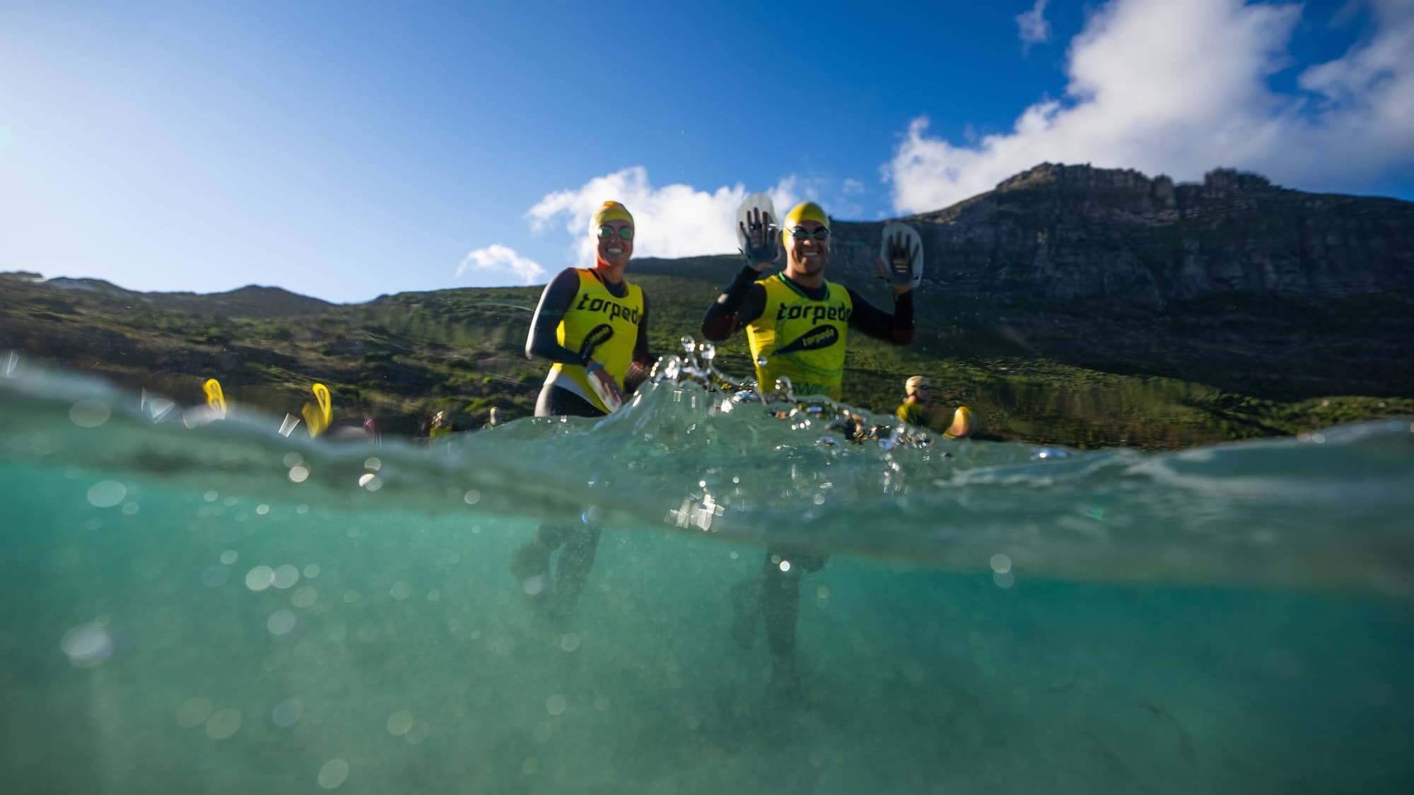 Two Torpedo athletes waving with hand paddles as they wade into the sea below the mountains