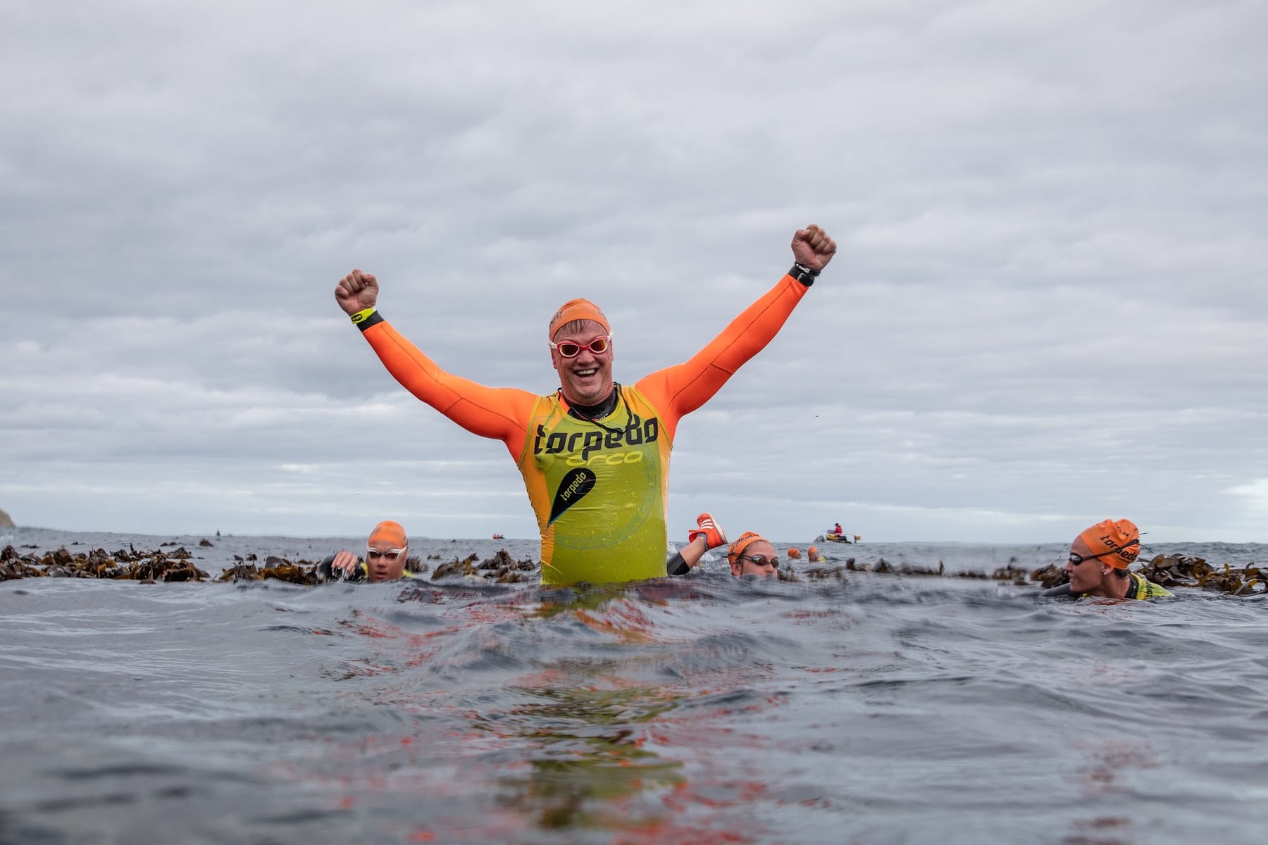 A swimmer in an orange wetsuit and yellow Torpedo vest throwing both arms up in the ocean, with other swimmers behind