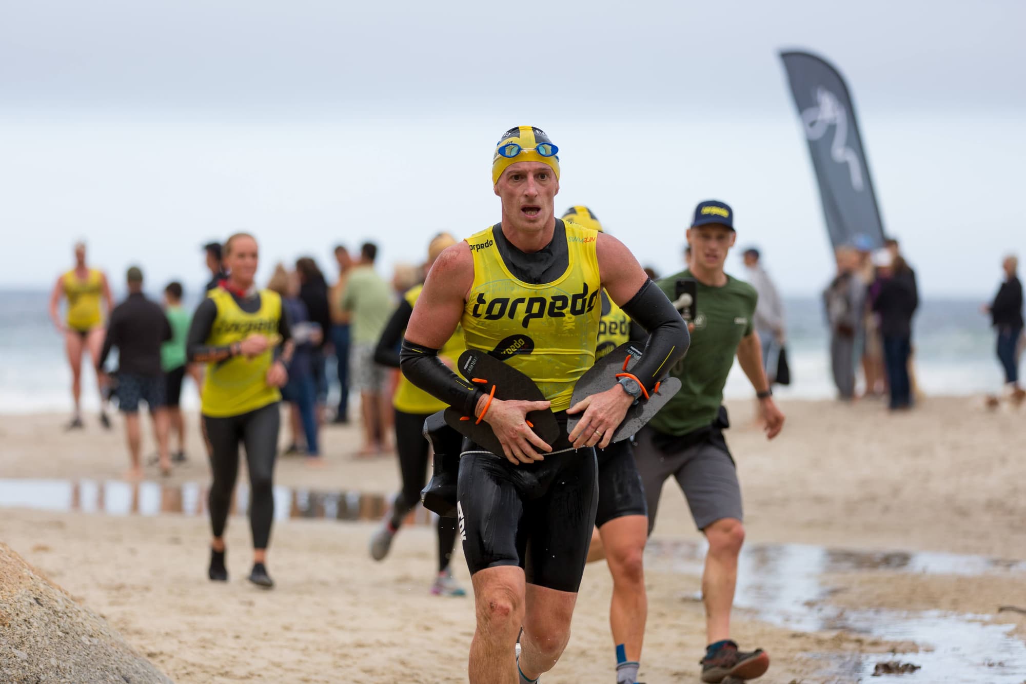 A swimrunner charging up the beach with hand paddles as teammates follow
