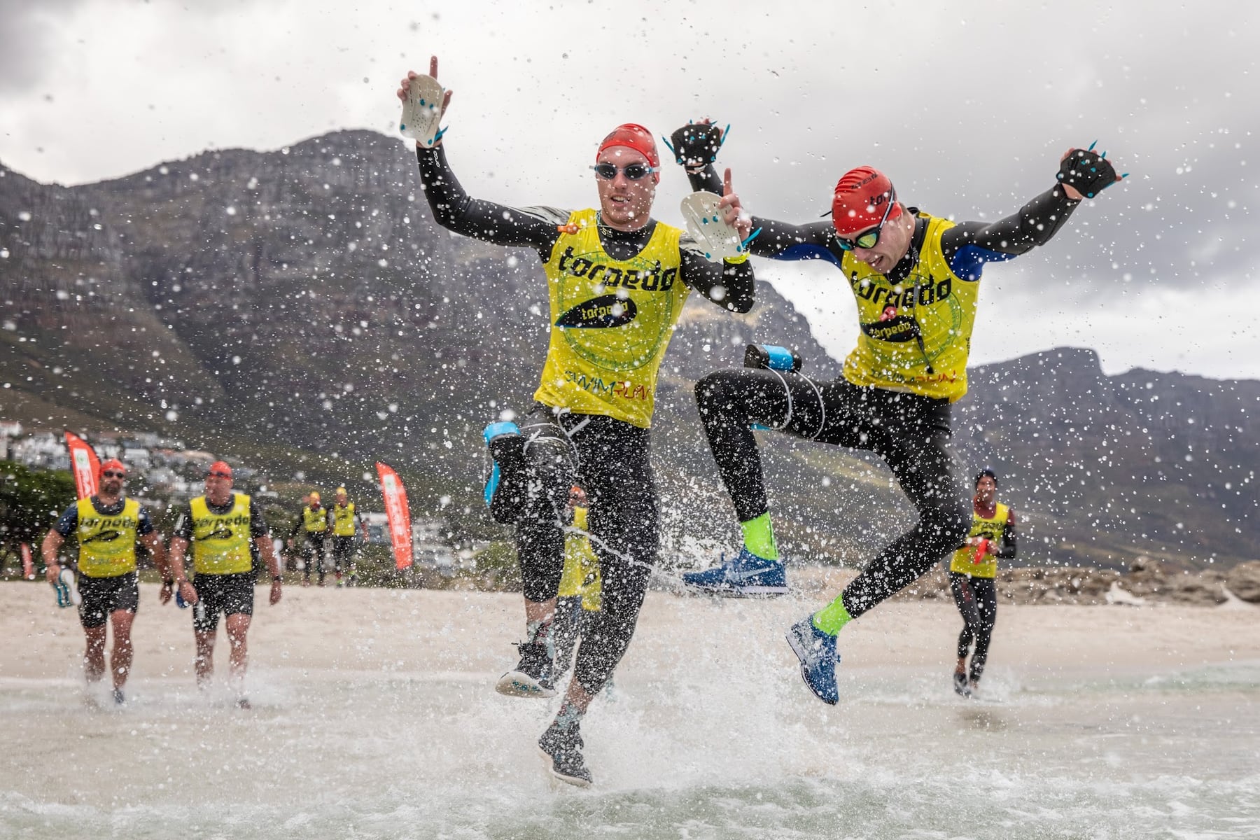 Two teammates in wetsuits and yellow vests leaping through the shallows with the mountains behind