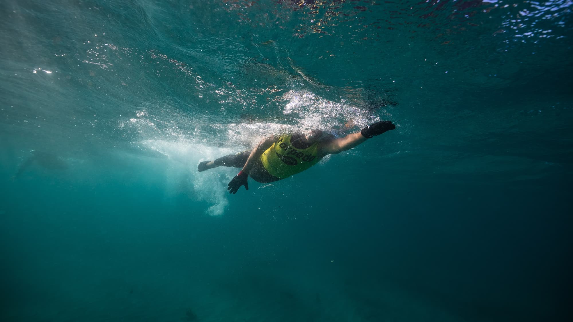 Underwater view of a gloved swimmer in a yellow Torpedo vest mid-stroke