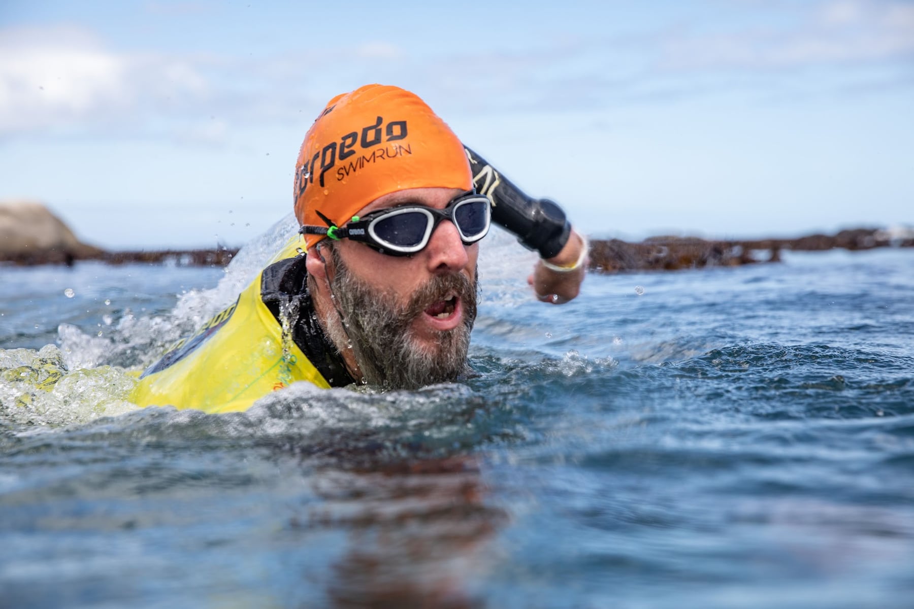 A bearded swimmer in an orange Torpedo cap and goggles swimming close to the camera