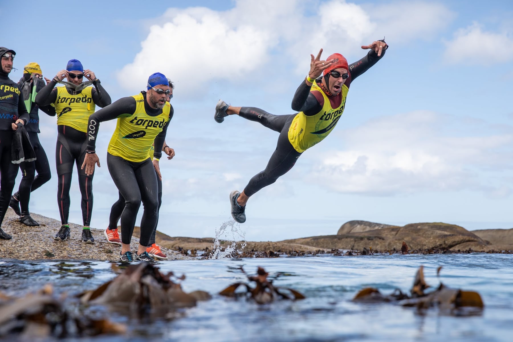 A swimrunner diving off the rocks into the kelp-filled sea while teammates watch