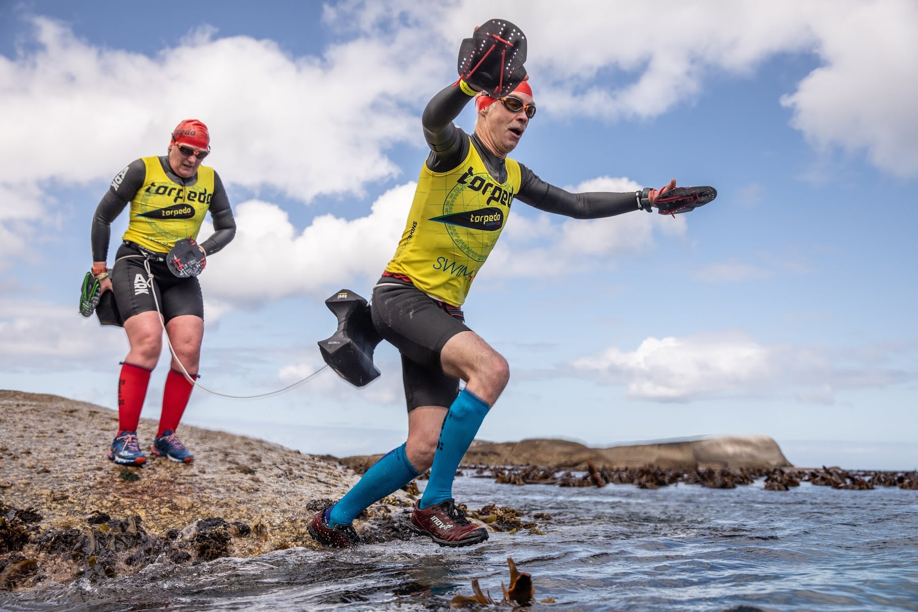 Two athletes with hand paddles and pull buoys running across coastal rocks