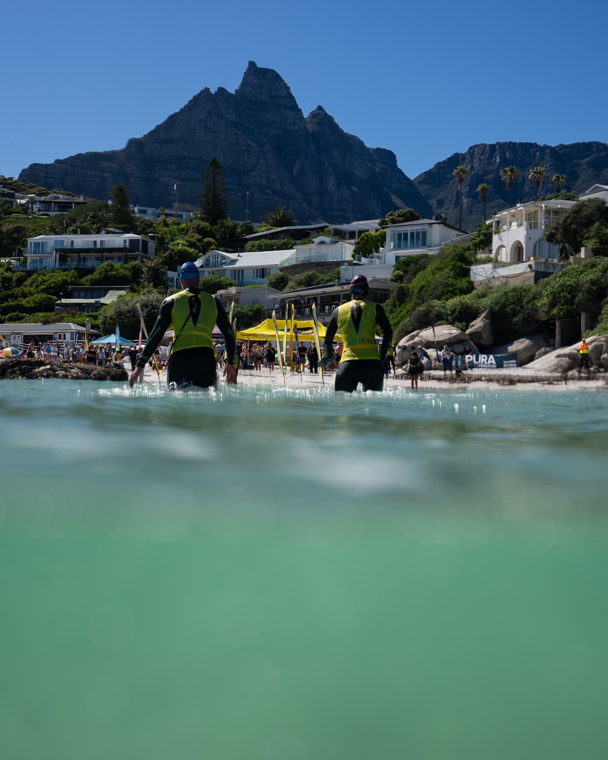 Two swimrunners wading towards the beach start with the Twelve Apostles and Table Mountain behind