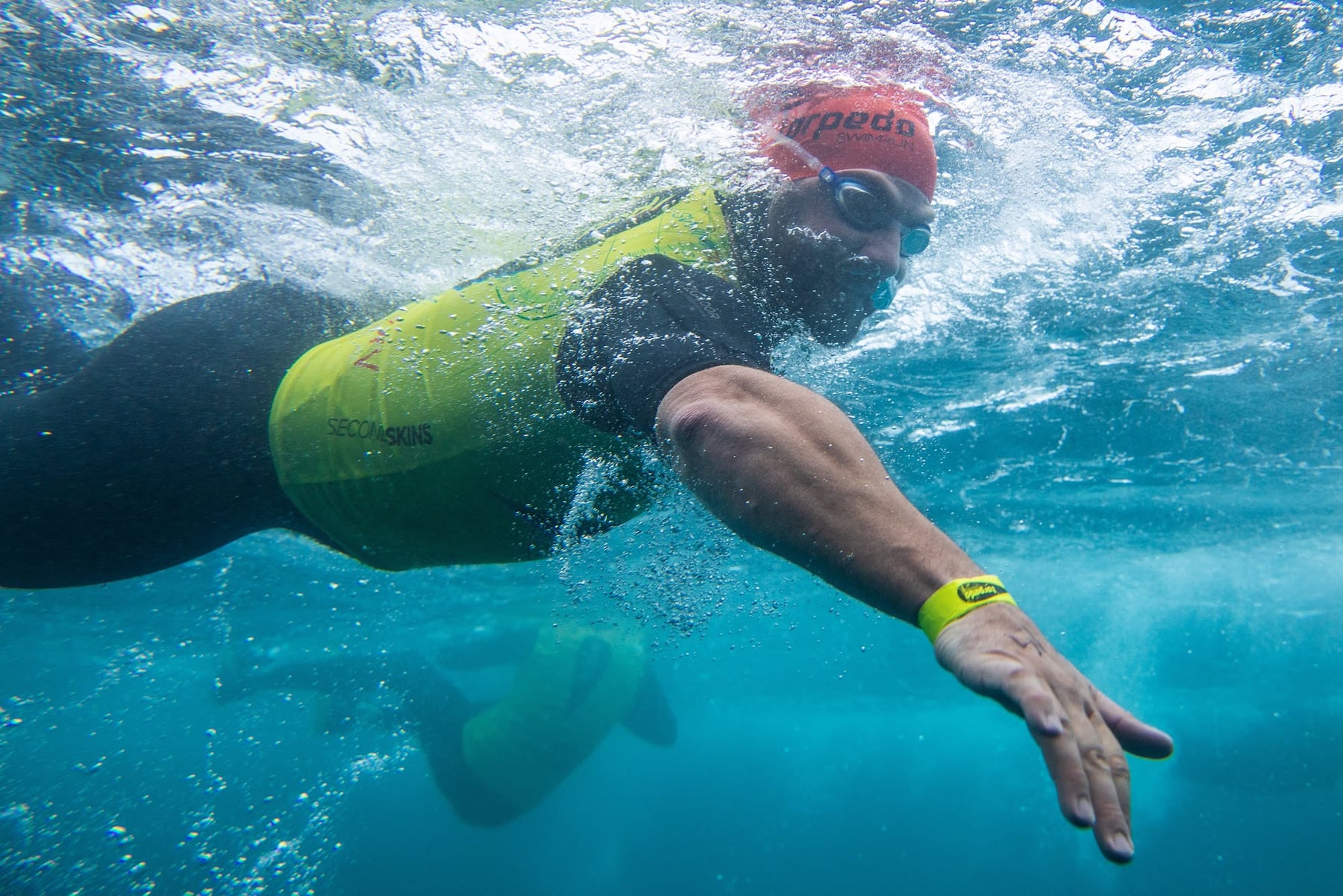 Half-underwater view of a swimmer in a red cap and yellow vest mid-stroke