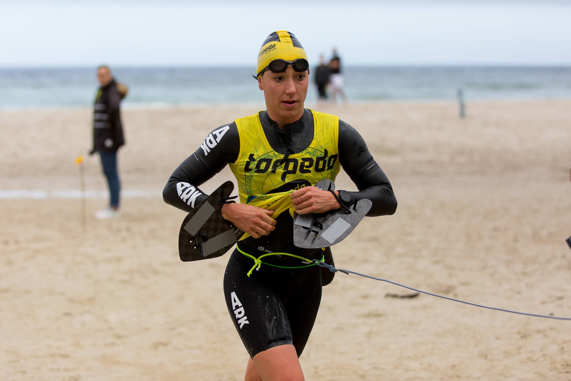 A swimrunner tethered to her teammate running up the beach with hand paddles