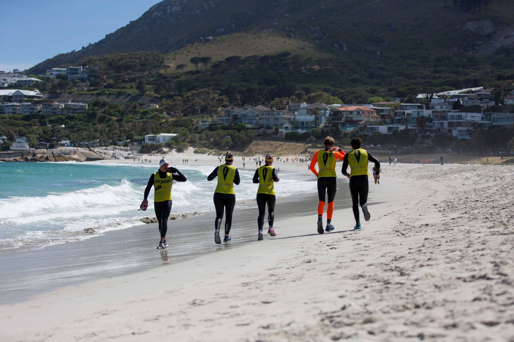 Five swimrunners in yellow vests running along Camps Bay beach beneath the mountain