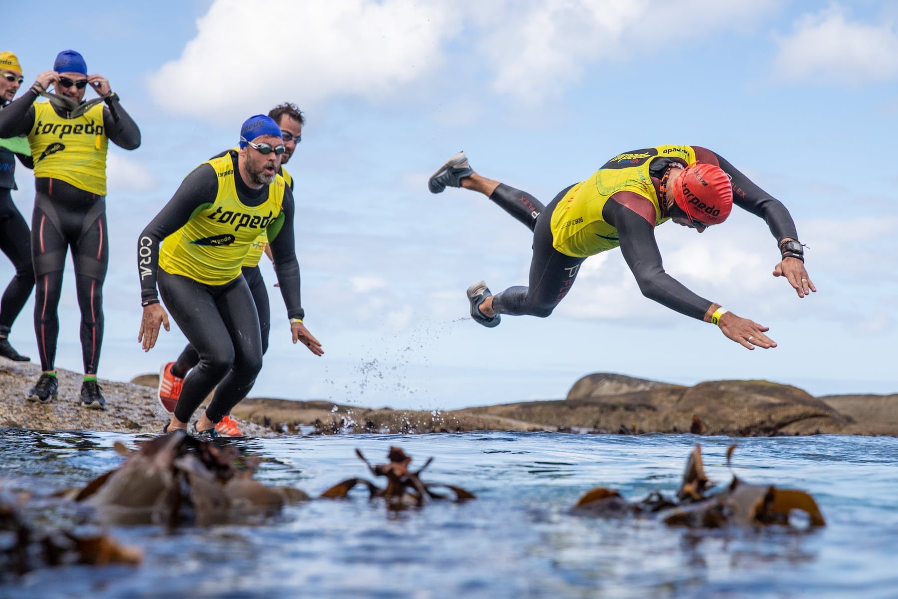 A swimrunner in a red cap diving from the rocks into the sea as teammates follow