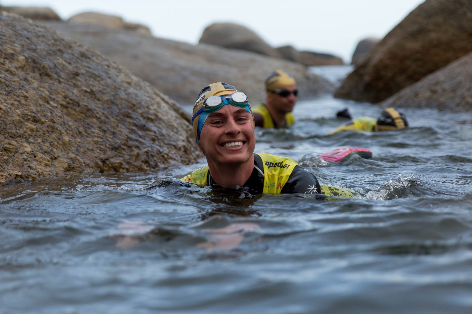 A smiling swimmer in a gold cap and goggles swimming through a gully between granite boulders