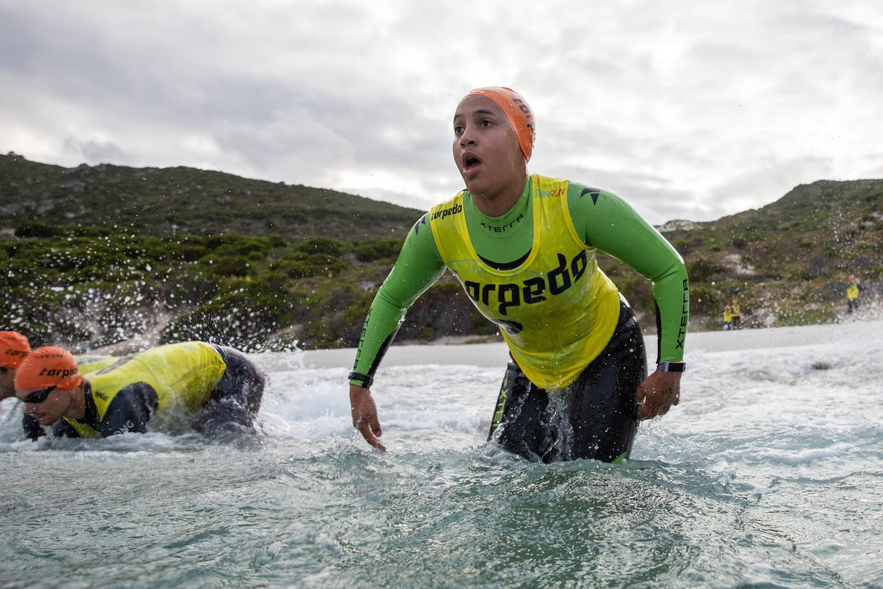 A swimmer in an orange cap and green-sleeved wetsuit wading out of the sea