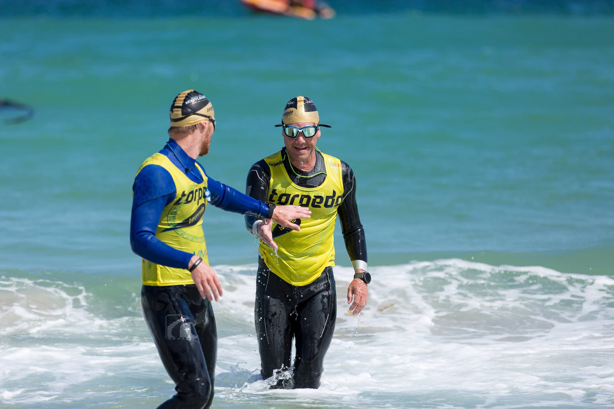 Two teammates in gold caps laughing together as they wade out of the surf