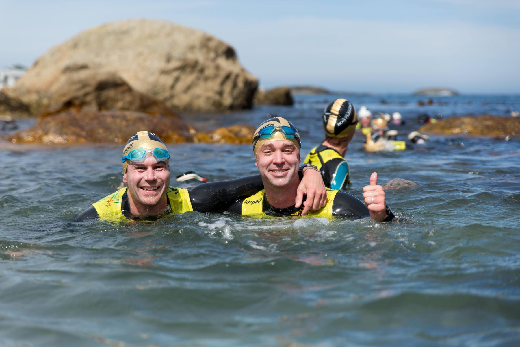 Two swimrunners in gold caps arm in arm in the sea, one giving a thumbs up