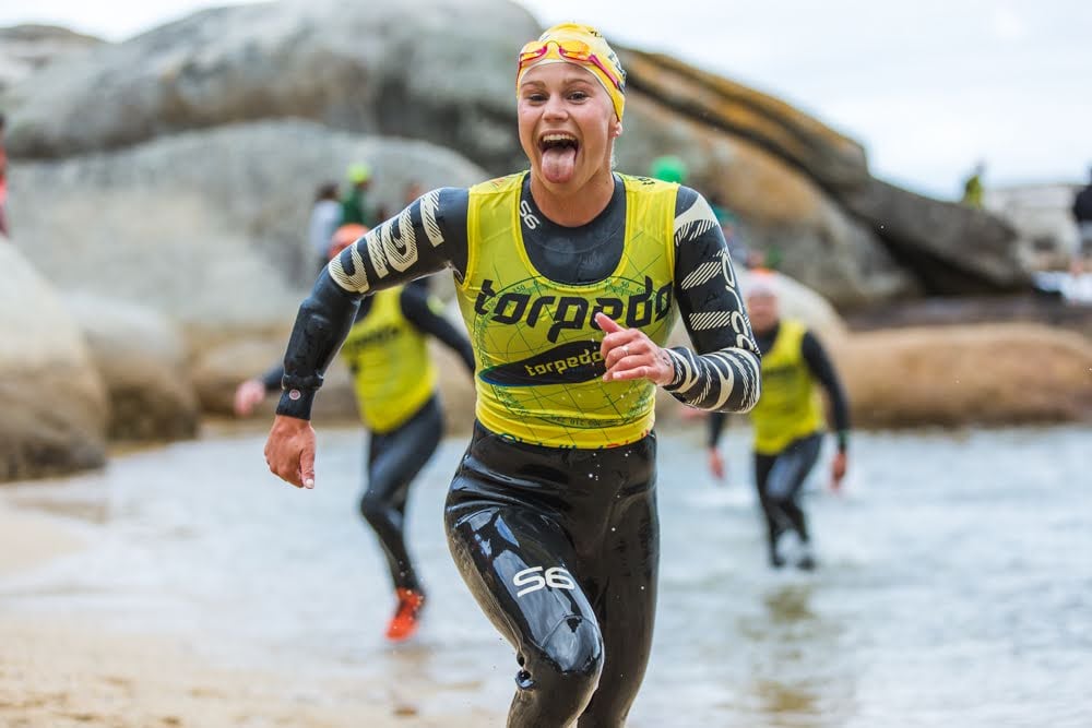 A grinning swimrunner in a yellow vest running out of the water past granite boulders