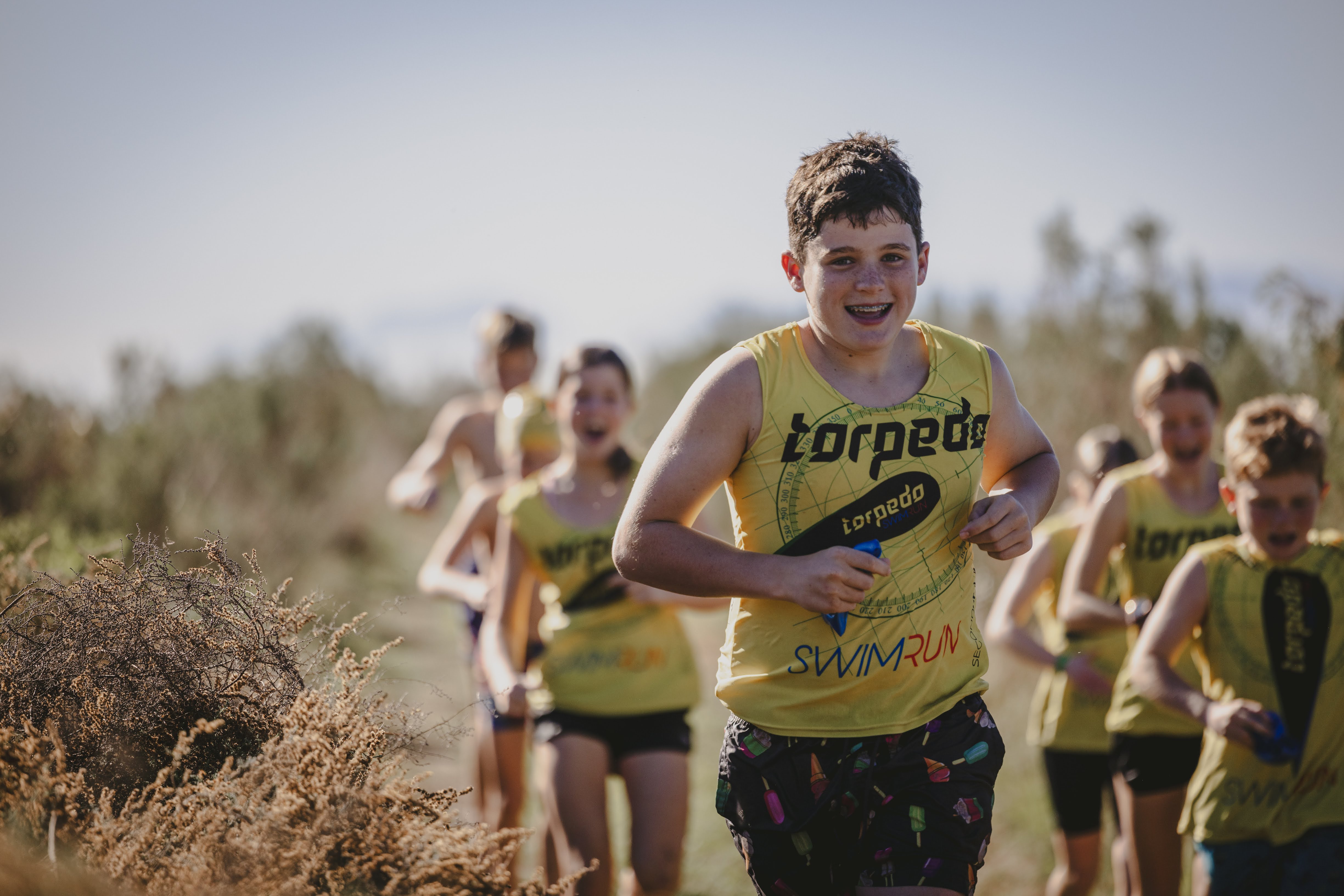A grinning boy in a yellow Torpedo vest leading a running pack through the fynbos