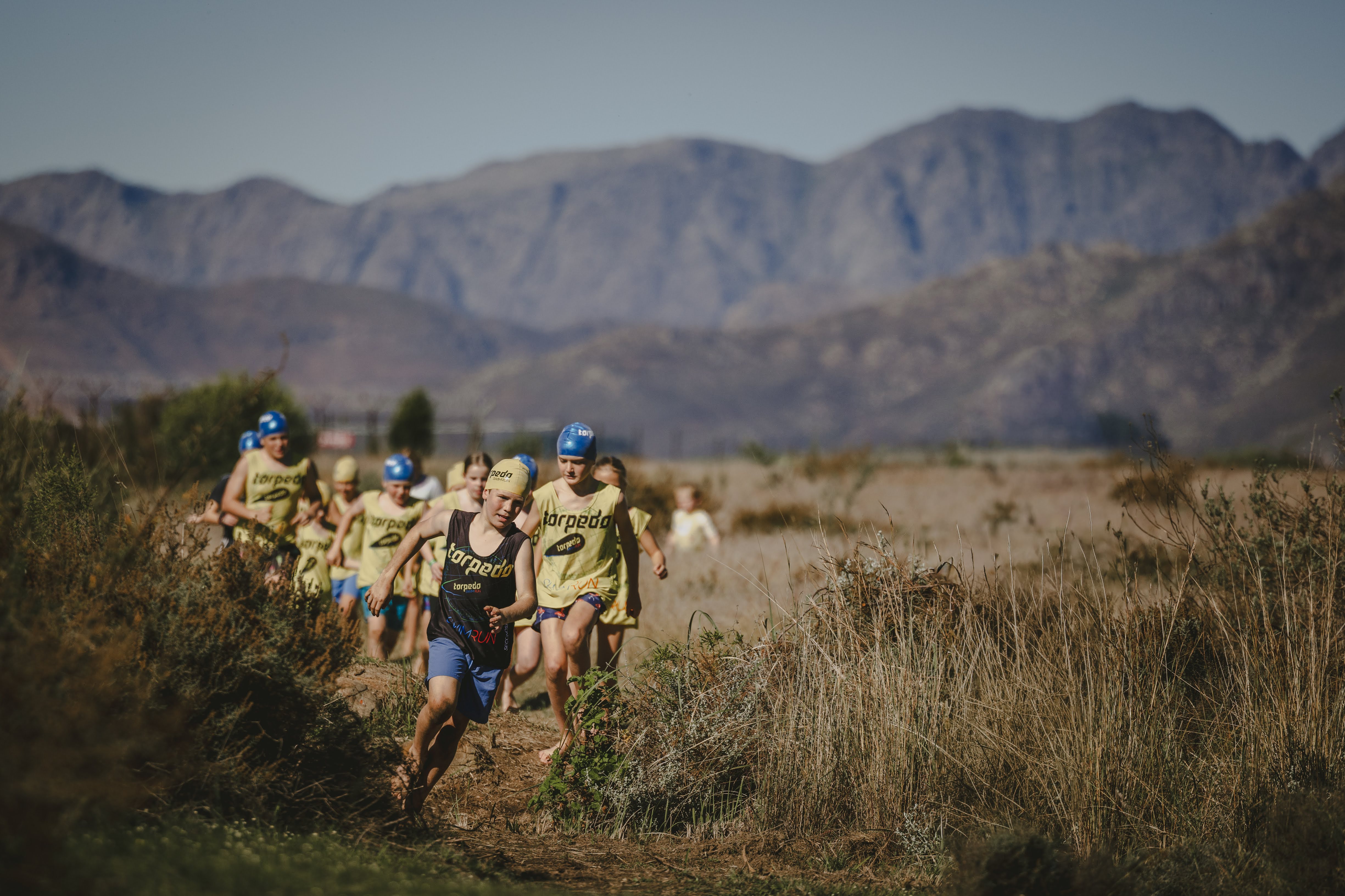 A boy in a black Torpedo vest leading a pack of kids running through the fynbos with mountains behind