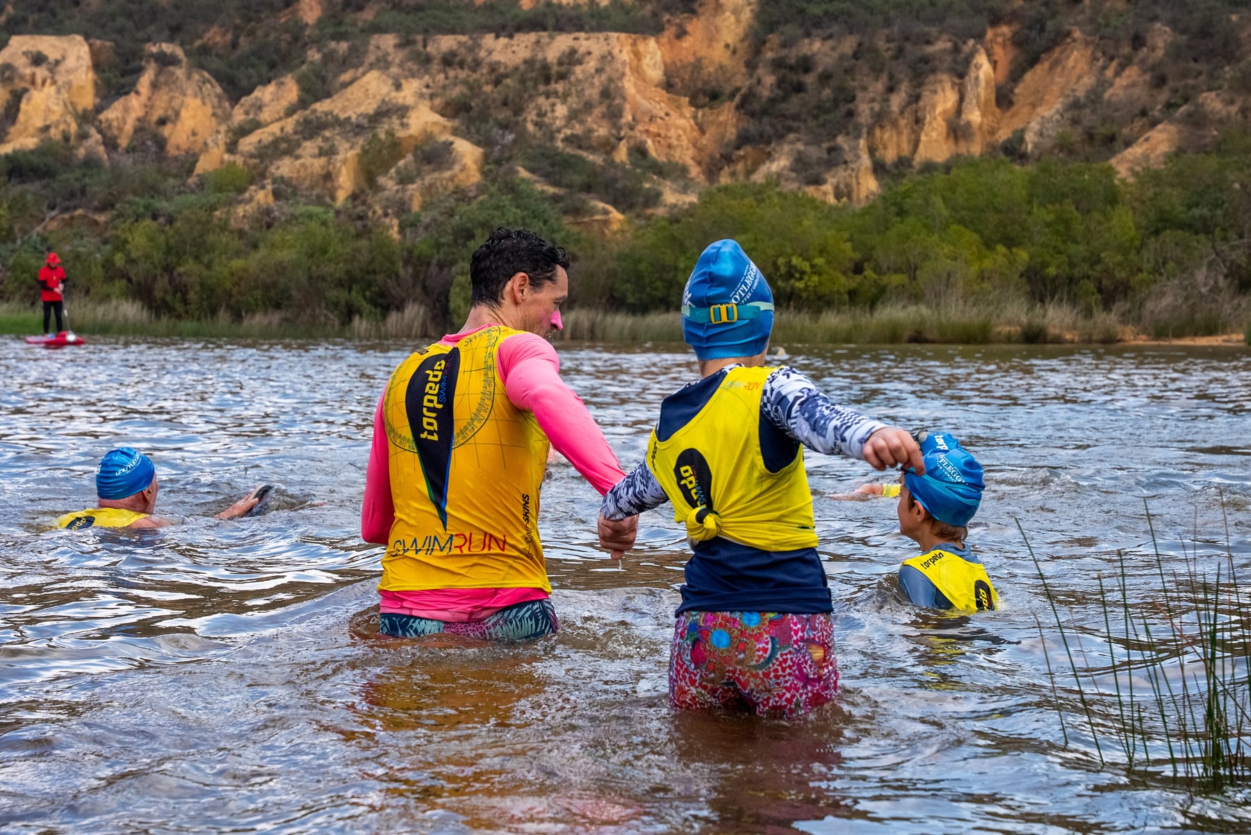 A dad and child holding hands as they wade into the river beneath sandstone cliffs, with other kids swimming alongside