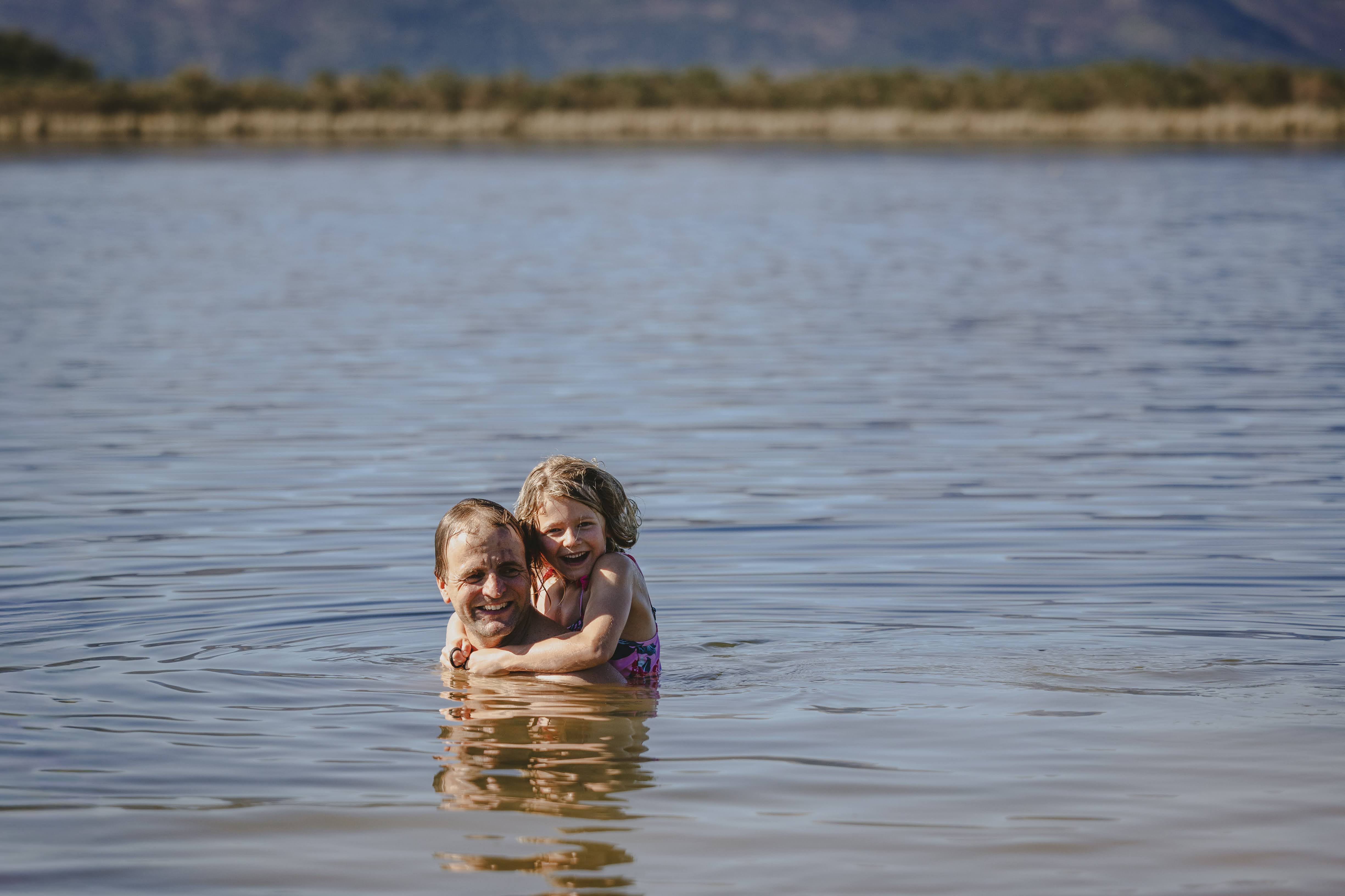 A dad in the dam with his young daughter hugging his neck, both laughing