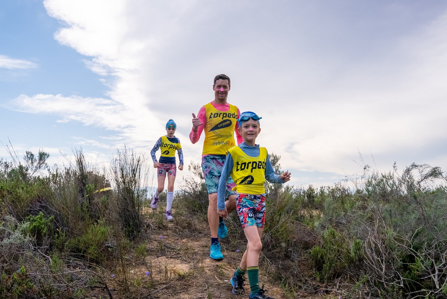 A dad giving a thumbs up as he runs down a fynbos trail with two kids in yellow Torpedo bibs