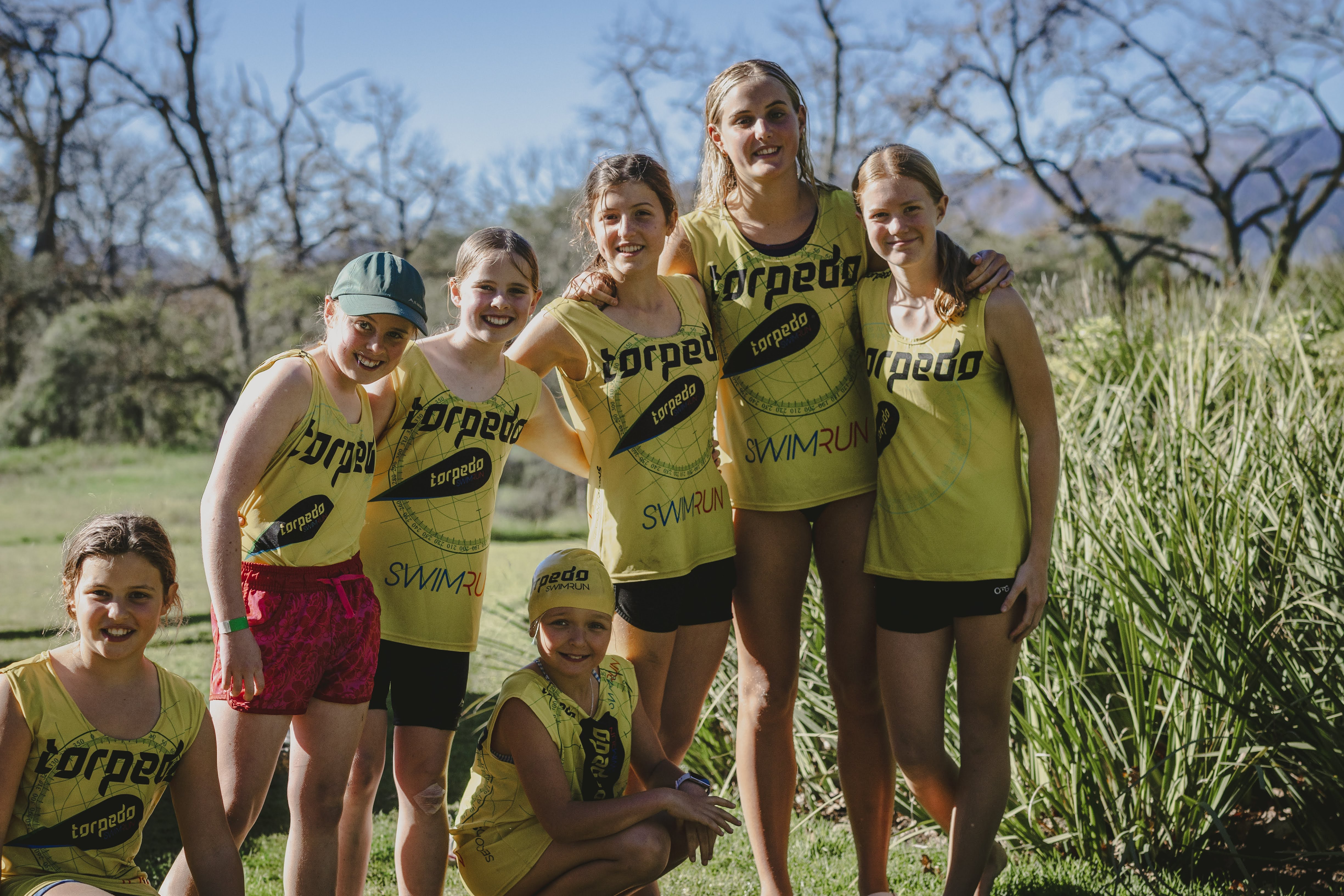 A group of seven girls in yellow Torpedo vests with arms around each other, smiling after their race