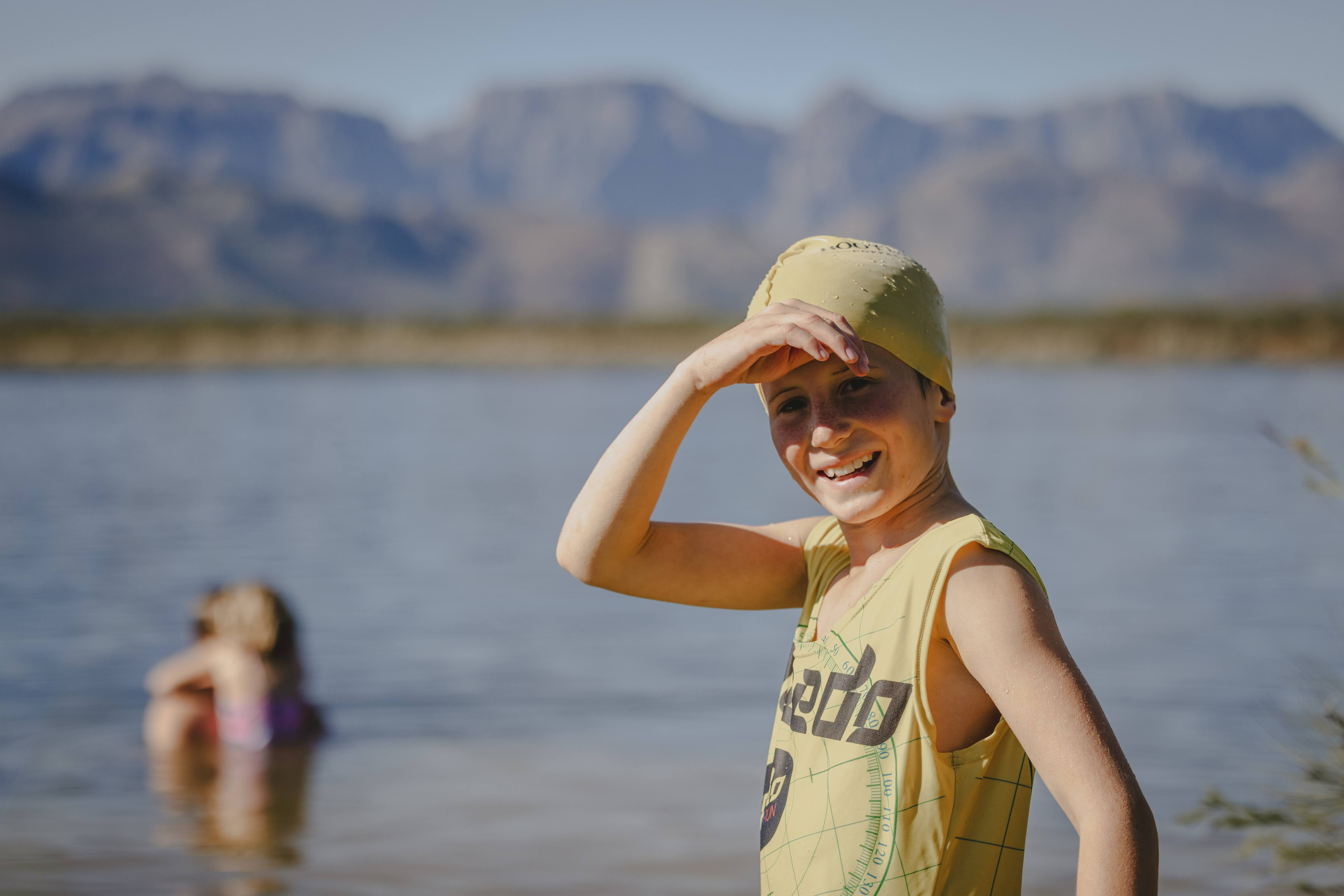 A grinning young swimmer in a yellow cap shielding their eyes from the sun at Plaisir dam