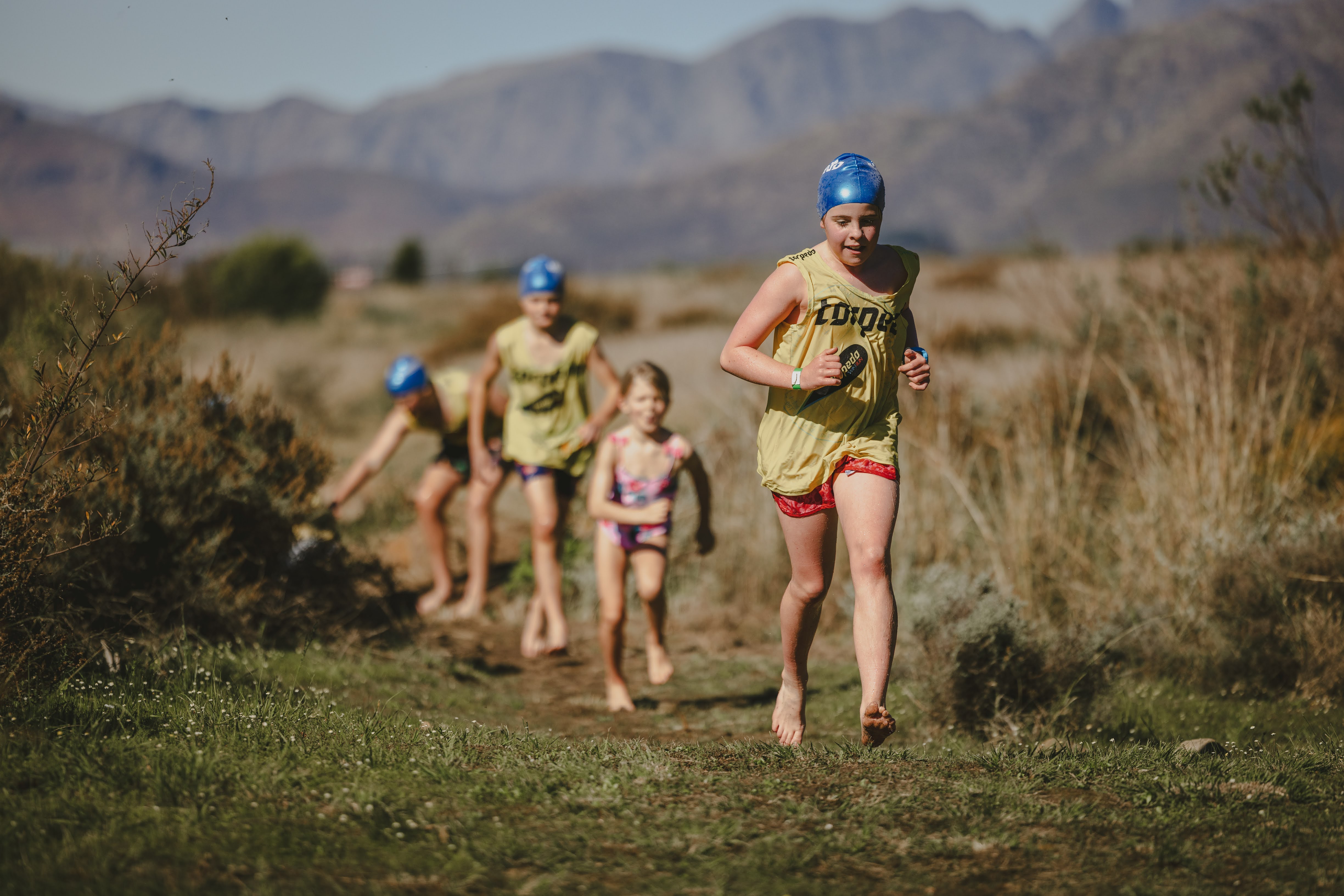 Four kids in caps and vests running toward the camera through the fynbos with mountains behind