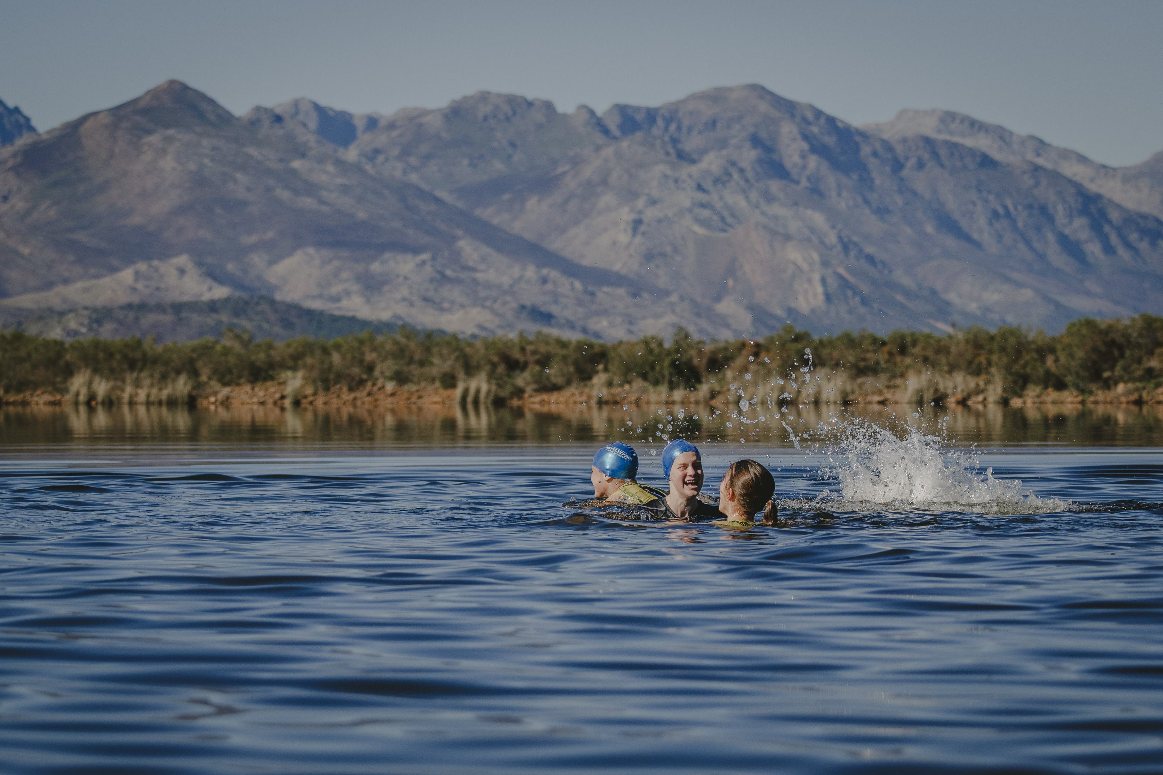 Three young Torpedo swimmers laughing and splashing in the dam with the Banhoek mountains behind them