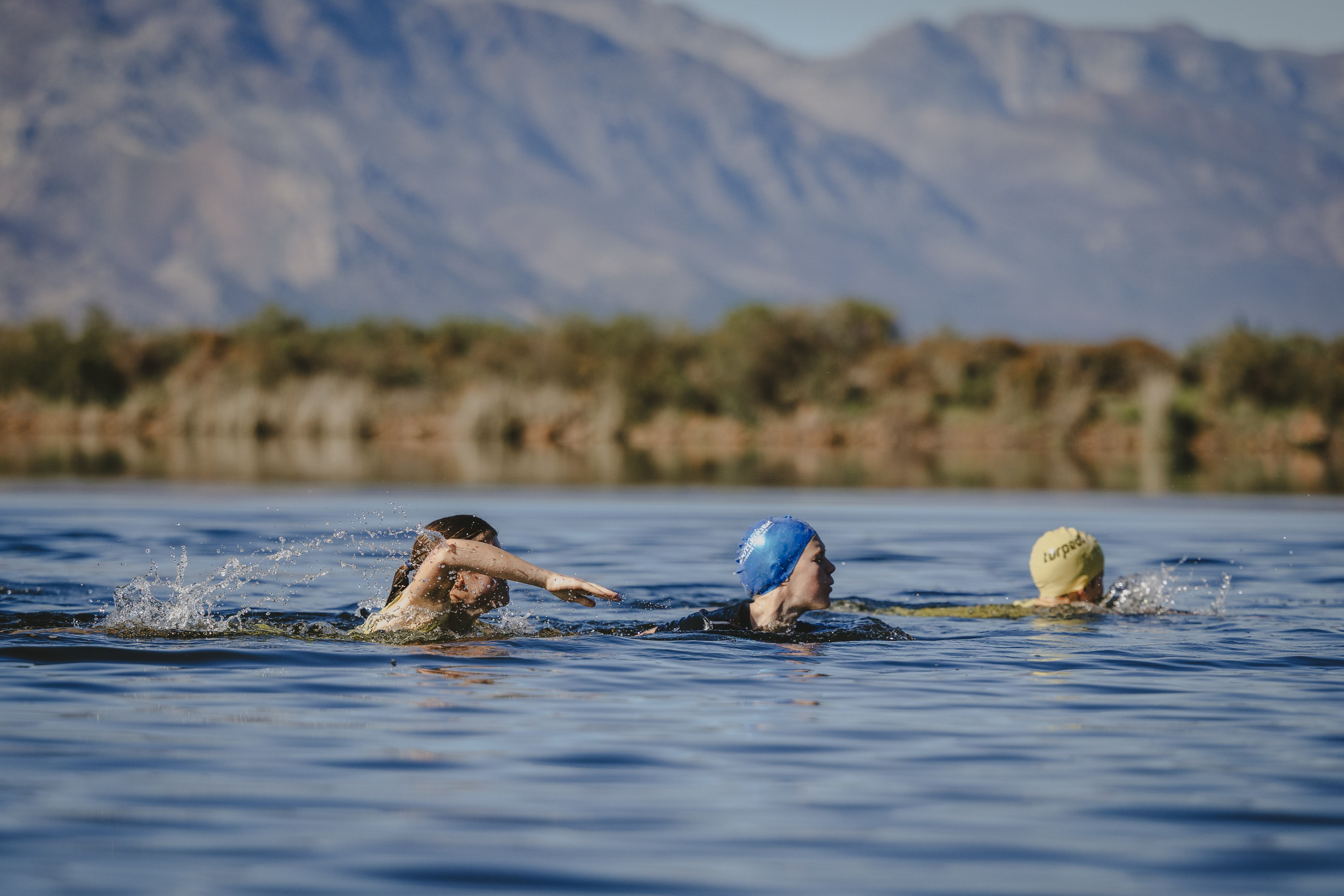 Three kids swimming across the dam, one mid-splash, with mountains behind