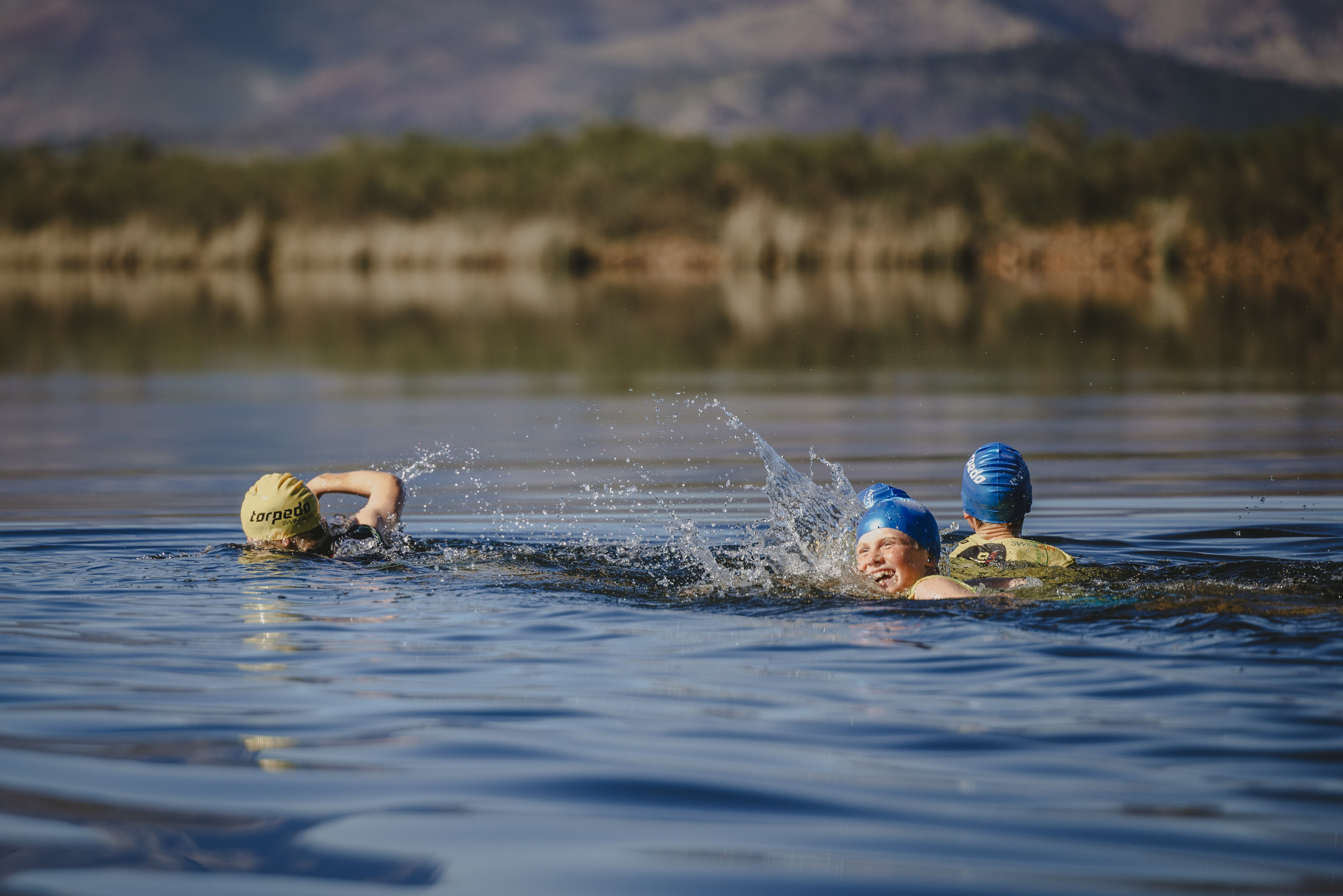 Two kids laughing mid-swim as a third throws up a big splash on the mountain-backed dam