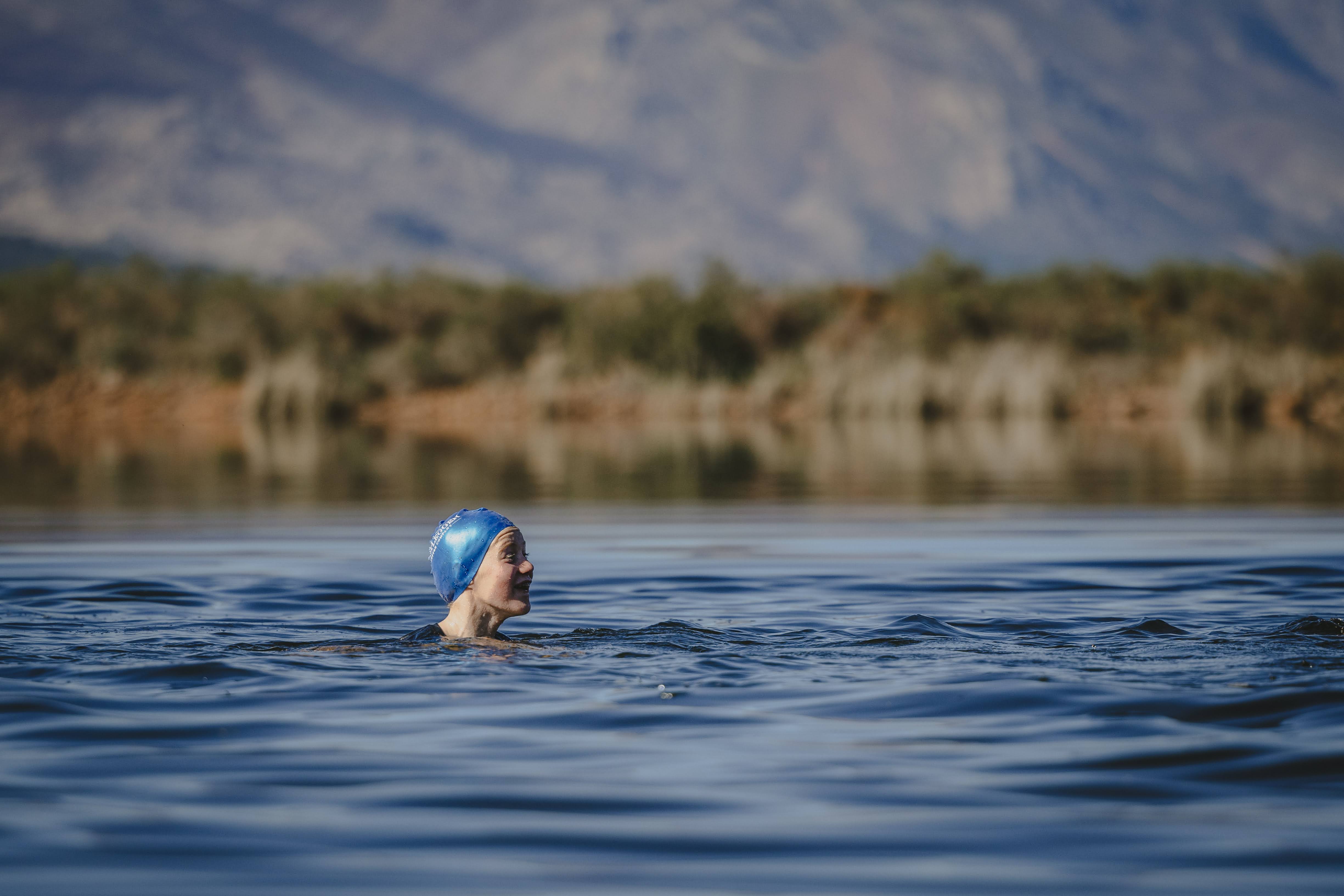 A solo young swimmer in a blue cap crossing the glassy Plaisir dam with mountain reflections