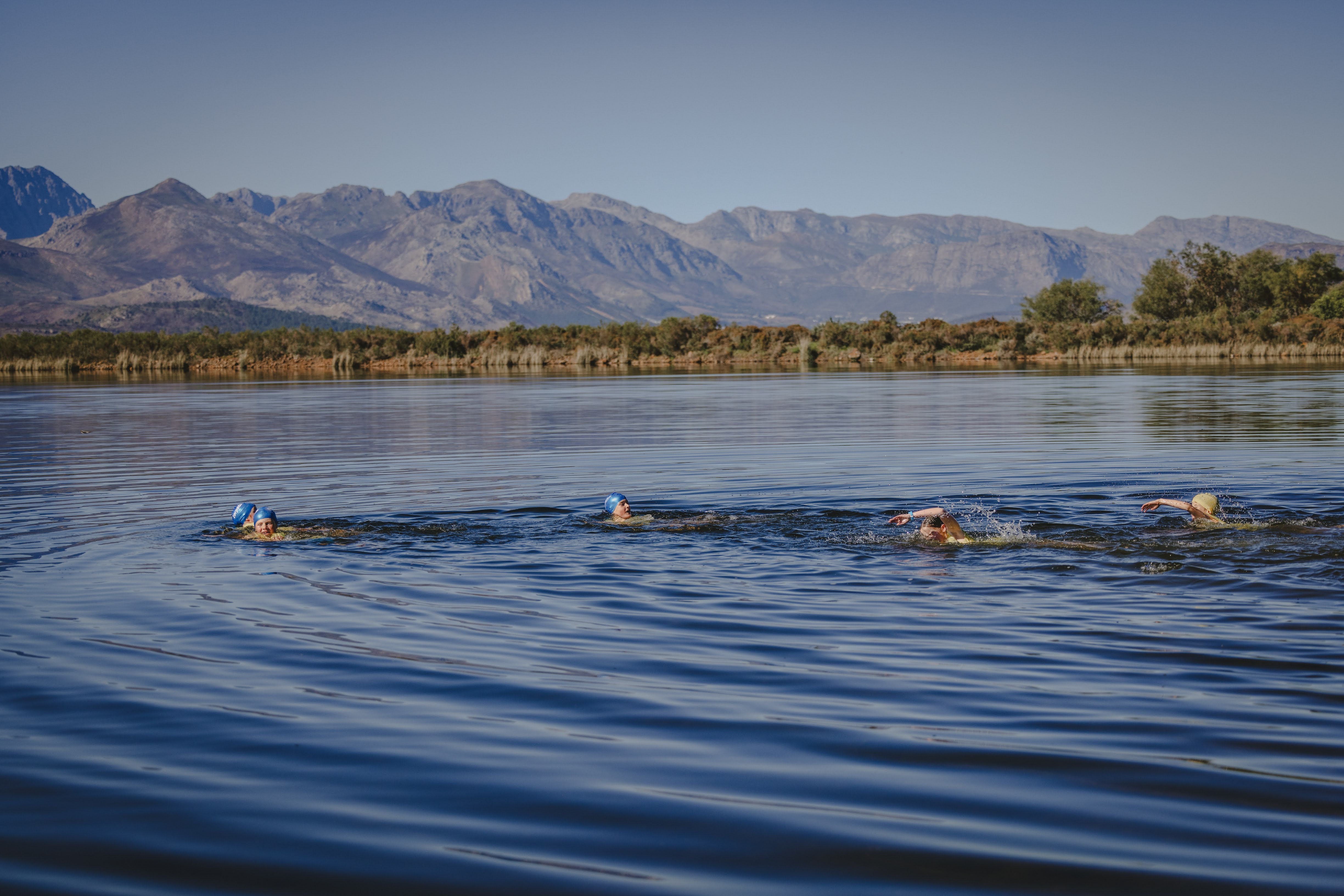 A line of young swimmers crossing the calm Plaisir dam beneath a sweeping mountain range