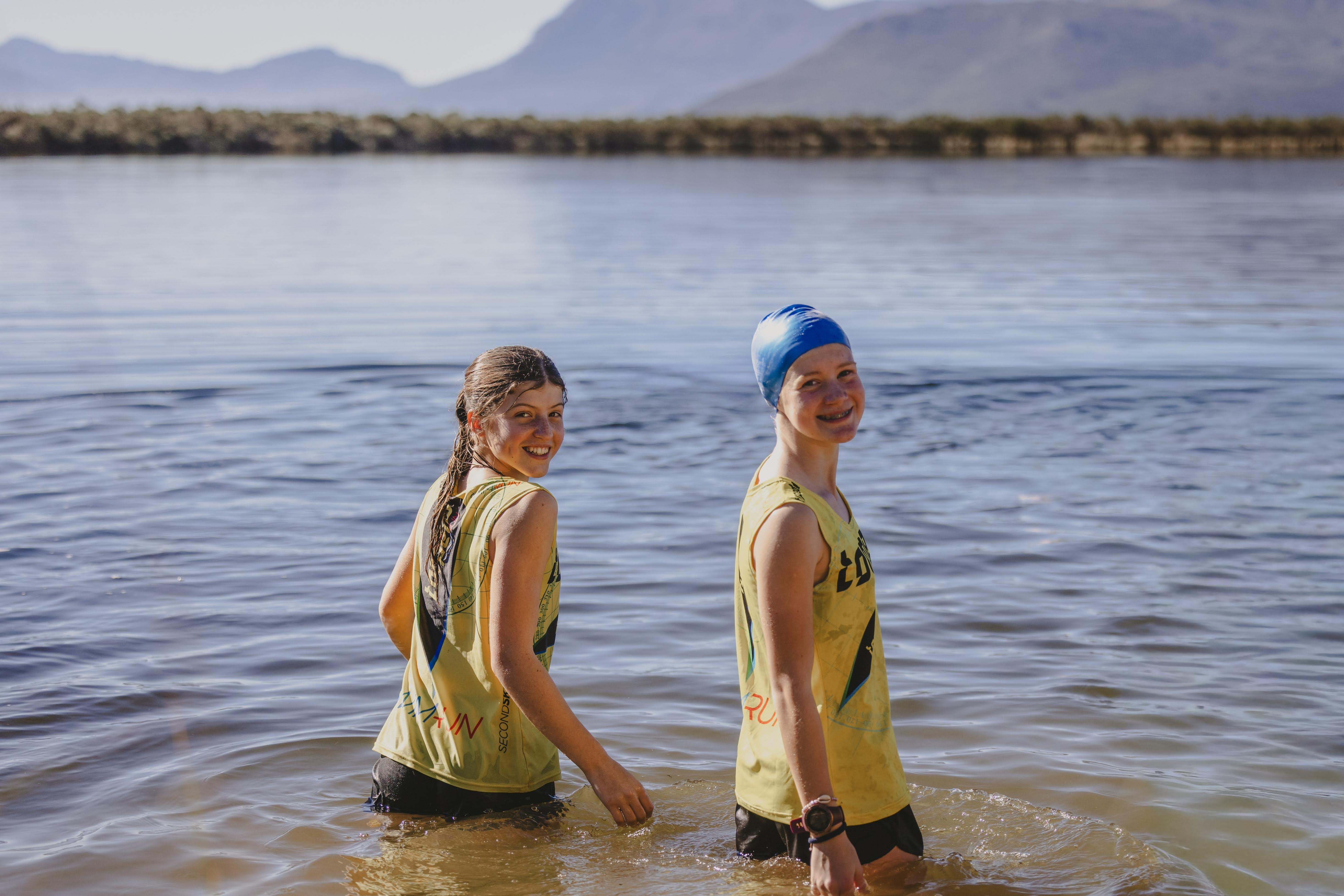 Two girls standing waist-deep in the dam, smiling back at the camera with mountains behind